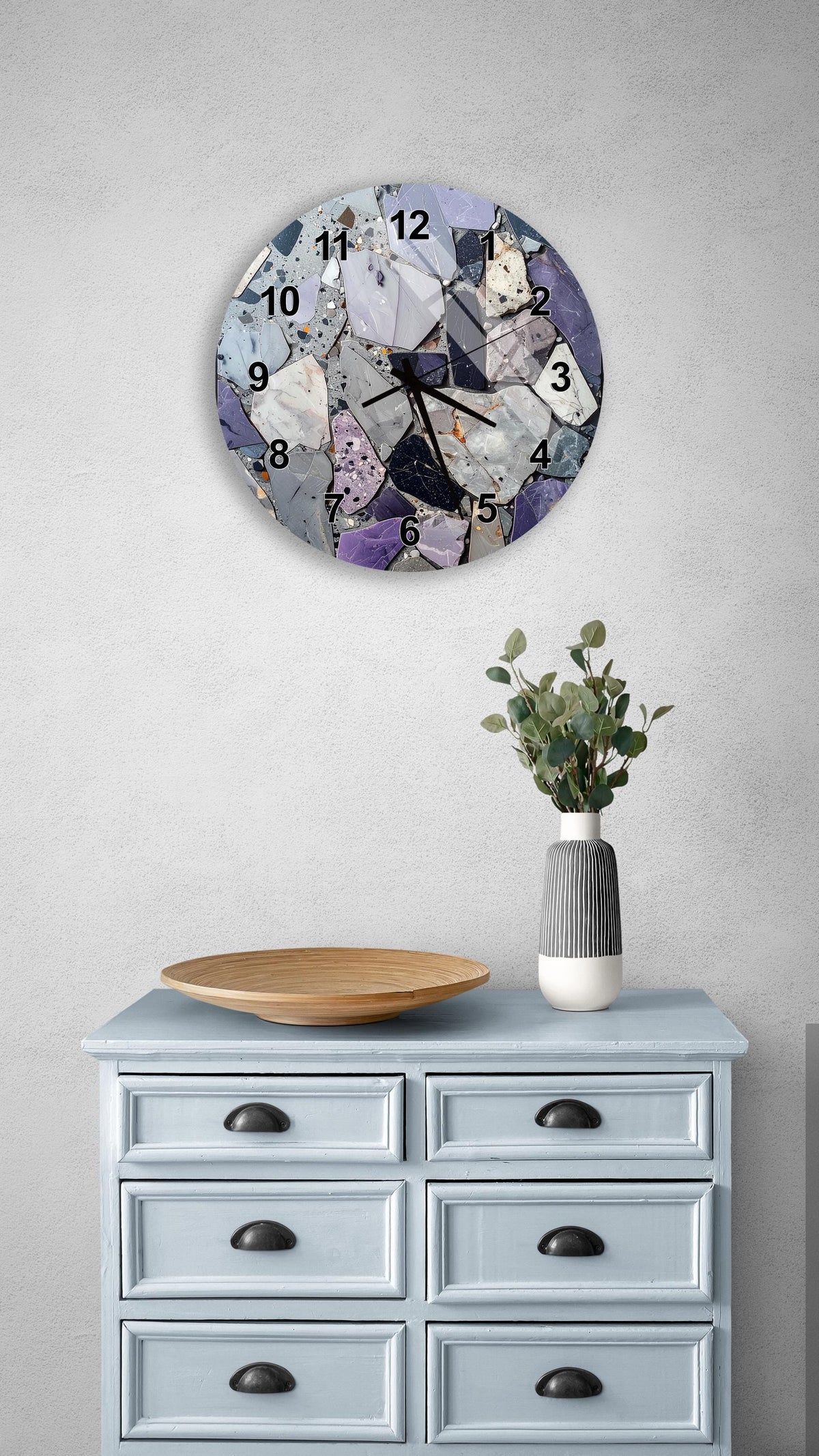 a clock with a mosaic design on a wall above a light blue dresser with six drawers. The dresser has a wooden tray on top and a vase with greenery on the surface.