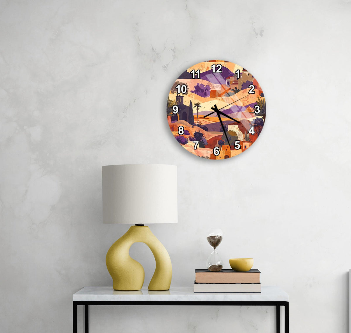 a round clock with a colorful, abstract design hanging on a wall above a white table with a yellow lamp and a stack of books.