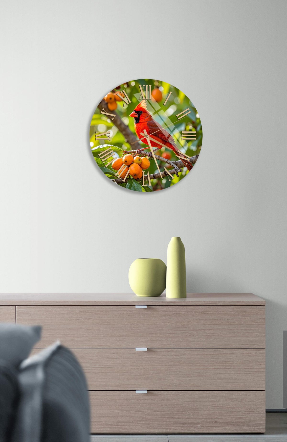 a round clock with a vibrant red cardinal perched on a branch, surrounded by orange berries, hanging on a white wall above a wooden dresser.