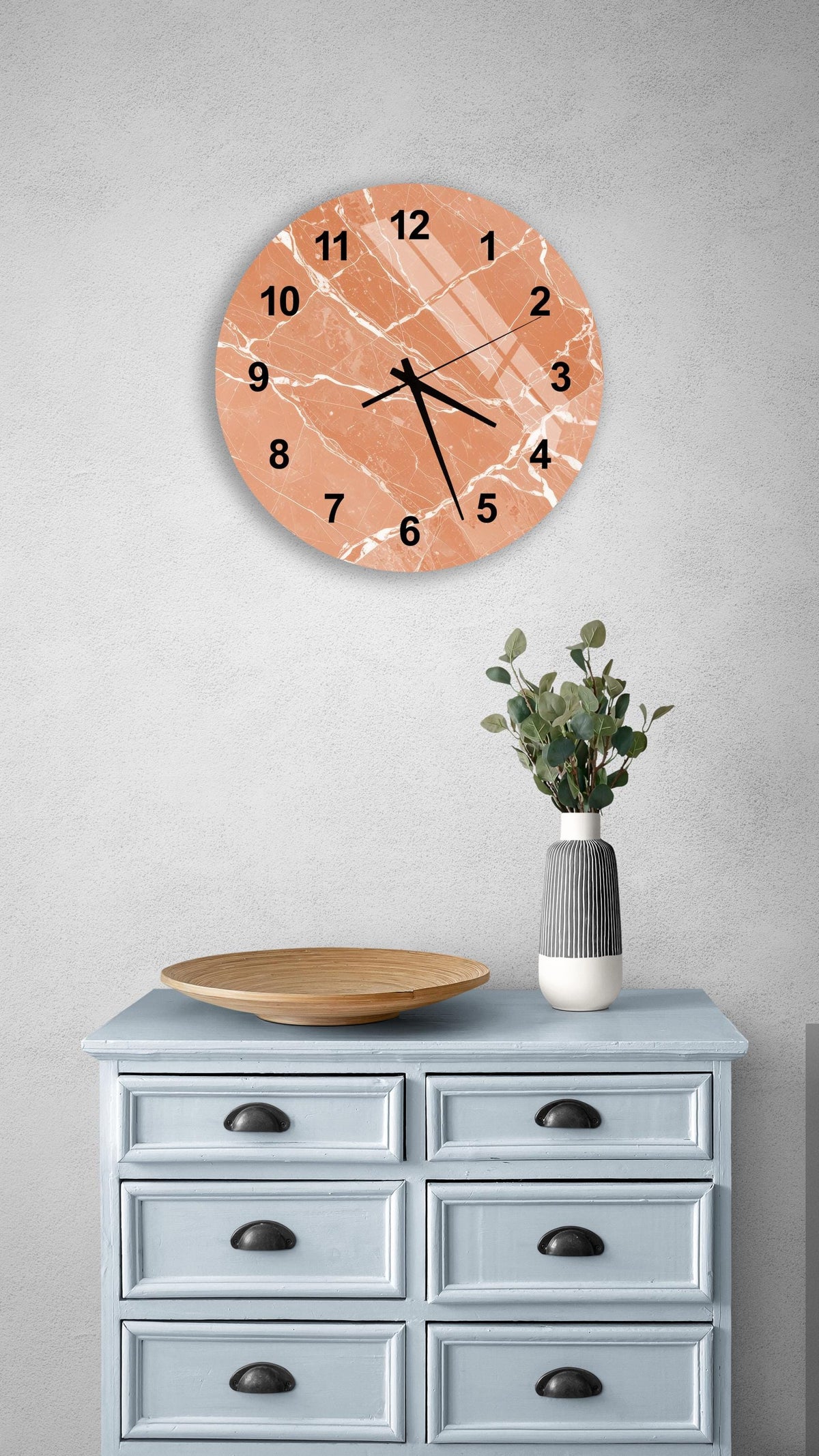 a wall clock with a marble-like design, mounted on a wall above a light blue dresser with six drawers. The dresser also has a vase with greenery on it, and there is a wooden plate on top of the dresser.