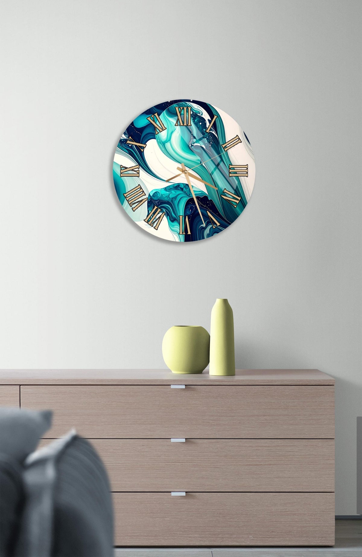 a modern clock with a turquoise and white design, hanging on a wall above a wooden dresser with three drawers. The dresser also has a small vase and a yellow bottle on top of it.