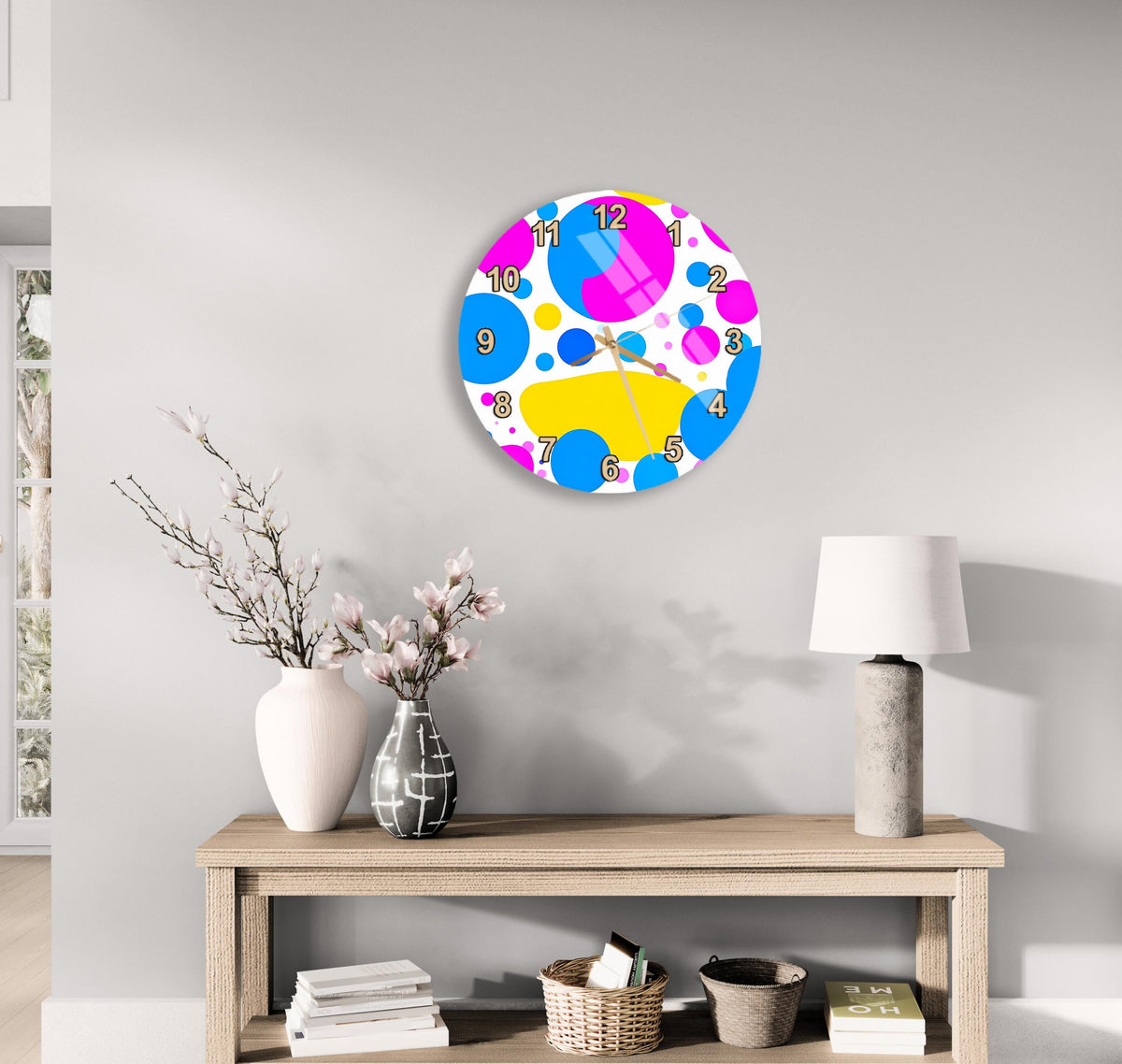 a colorful clock with a pattern of circles and dots on a white wall, accompanied by a wooden table with a vase of flowers, a lamp, and some books.