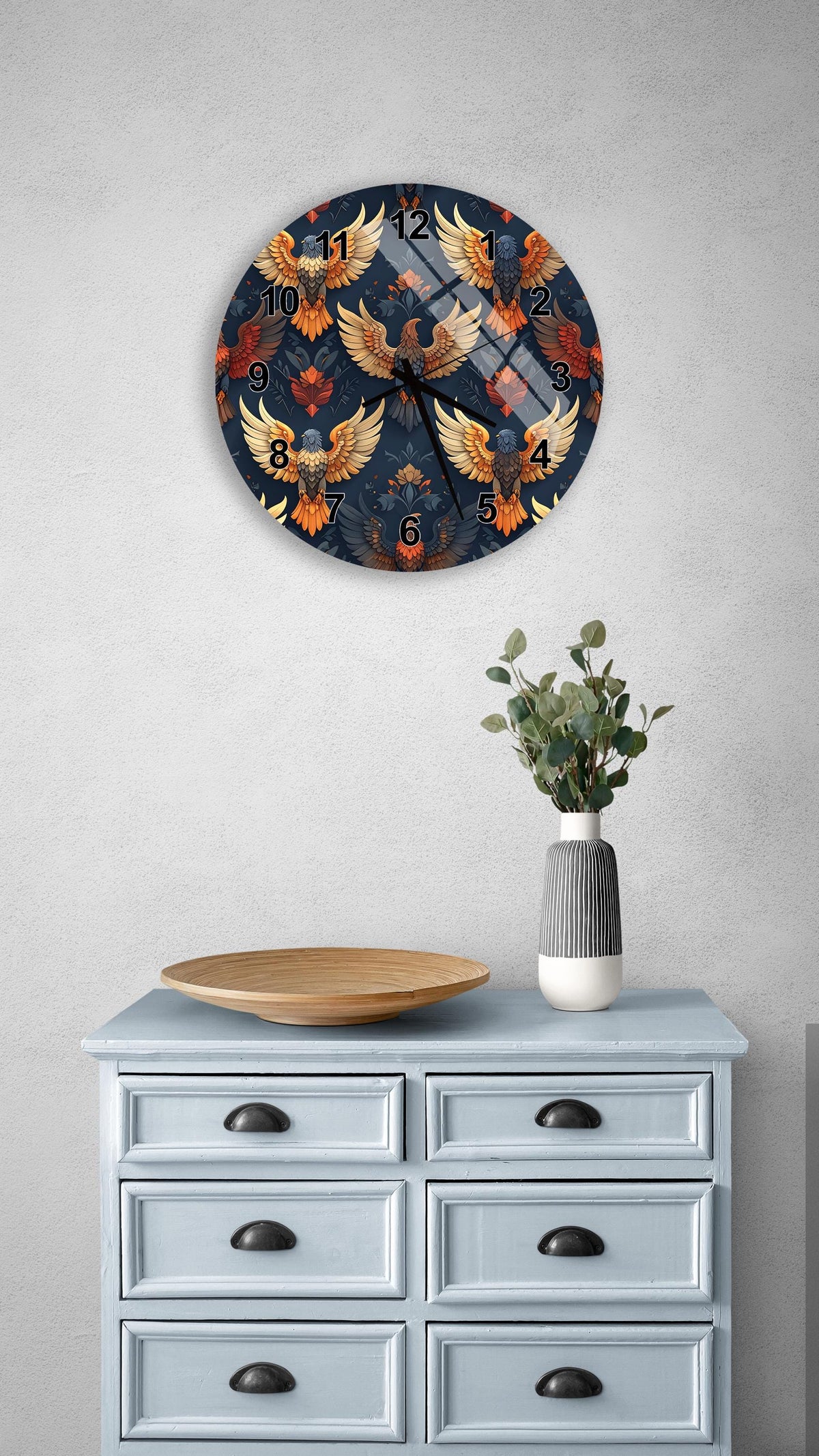 a wall clock with a vibrant, colorful design of birds and a vase with greenery on a light blue dresser.