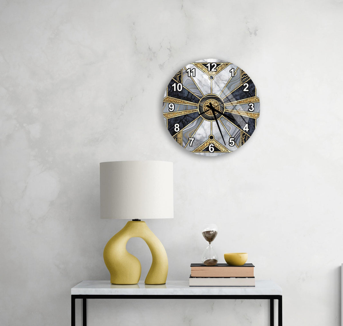 a modern clock with a unique design, mounted on a wall above a white table with a yellow lamp and a stack of books.