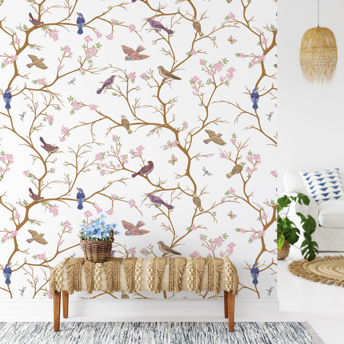 a wallpapered wall with a decorative pattern of birds and flowers, including blue birds and pink flowers. In front of the wall, there is a bench with a woven seat and a basket of flowers on it. The room also includes a white couch, a woven rug, and a potted plant.