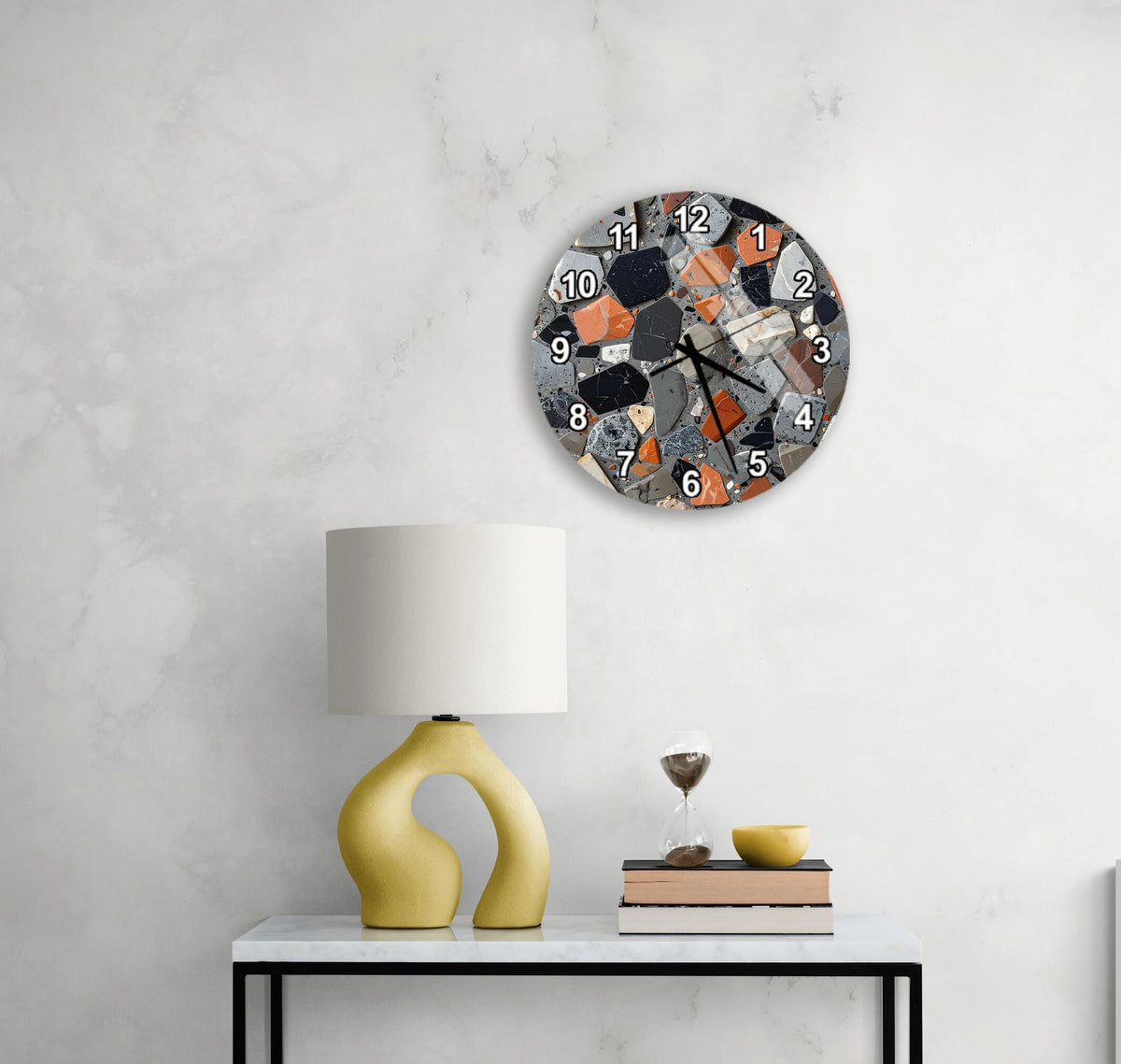 a modern, abstract-style clock with a mosaic design on the wall above a white table with a yellow lamp and a stack of books.