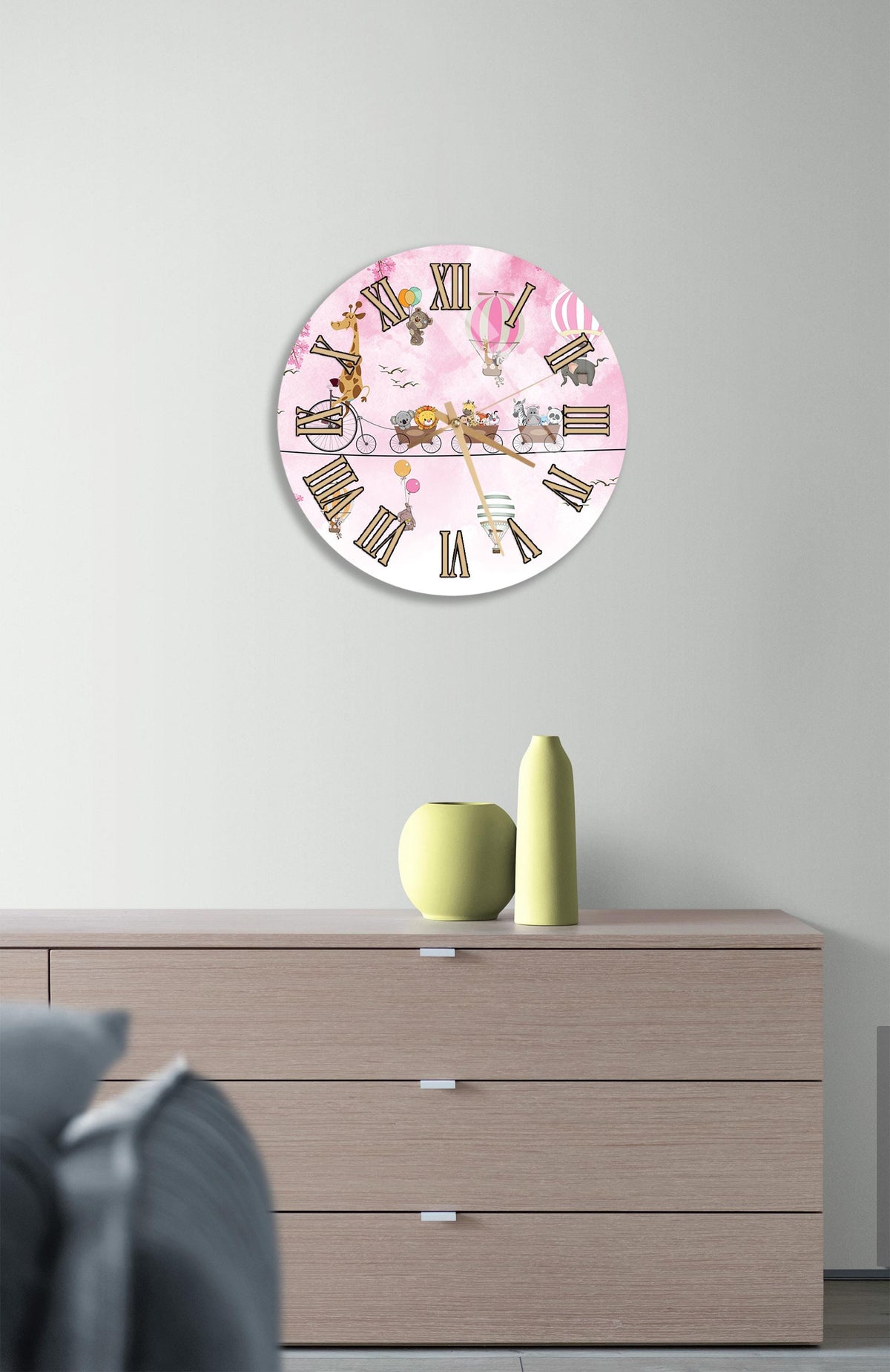 a round clock with a pink background and Roman numerals, hanging on a wall above a wooden dresser.