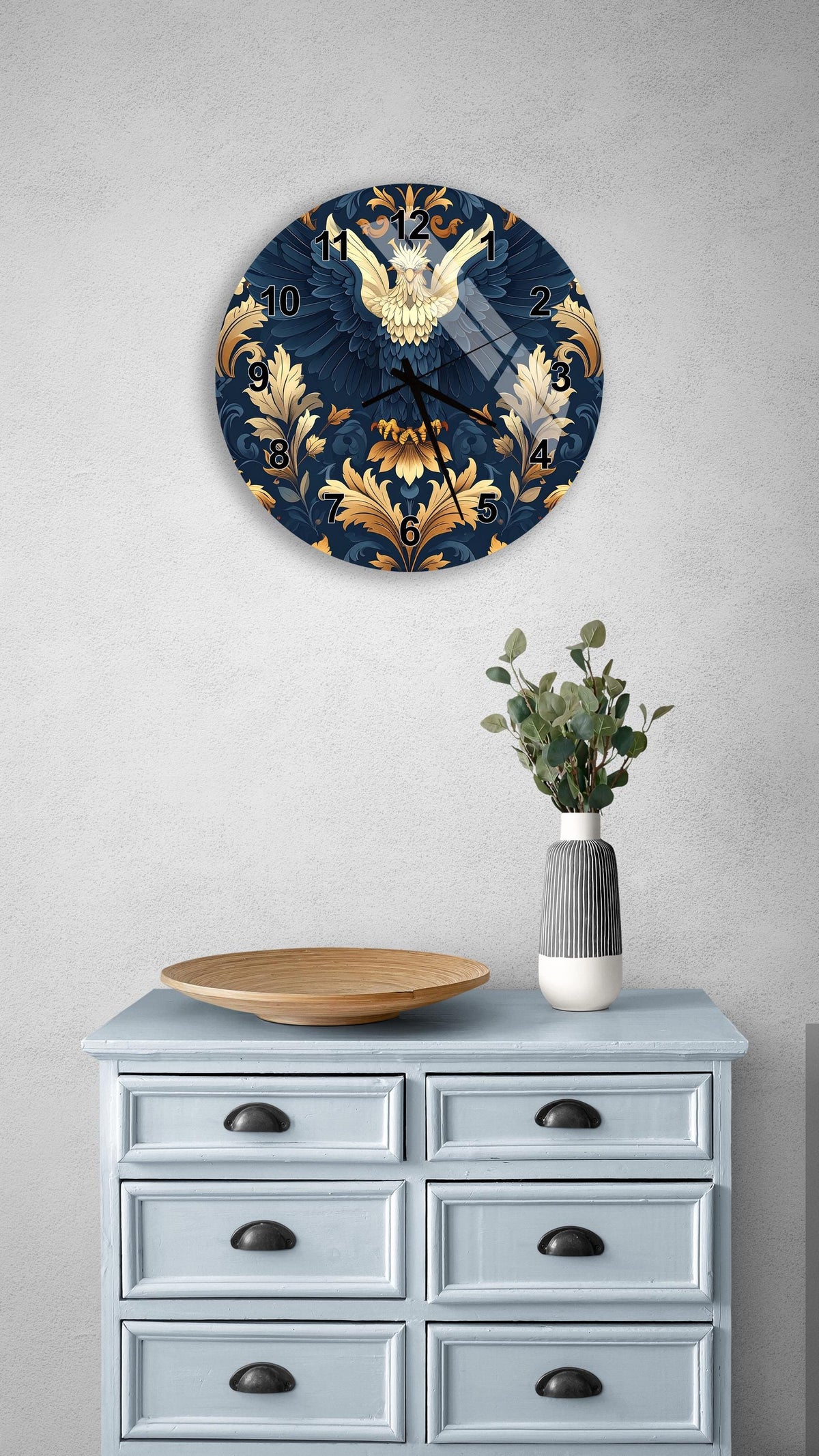a wall clock with a blue and gold design, mounted on a wall above a light blue dresser with six drawers. The dresser also has a vase with greenery and a wooden bowl on it.