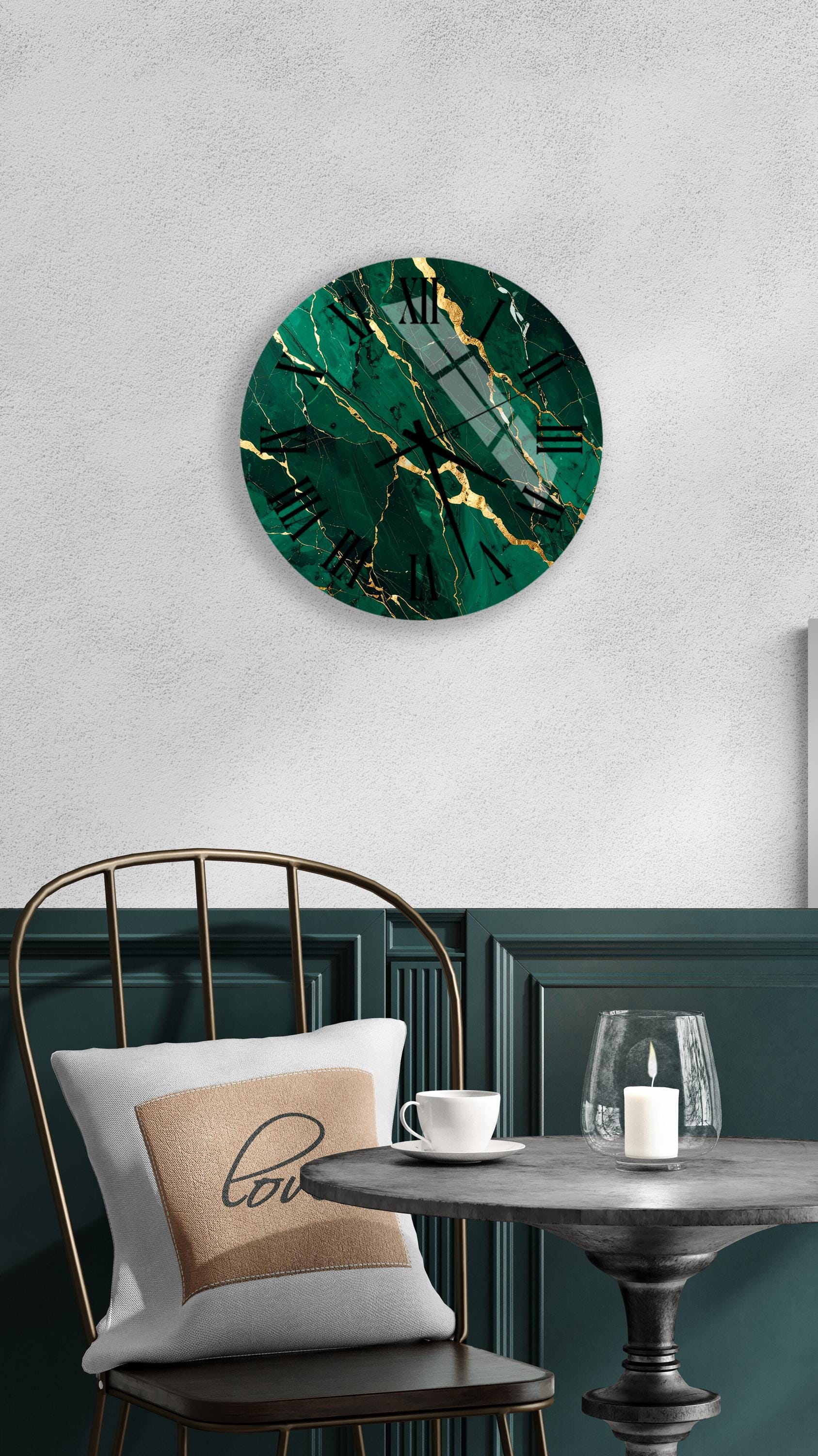 a modern, green-colored wall clock with a gold-colored design, hanging on a white wall above a black metal chair. The chair has a white pillow with the word "love" printed on it. In front of the chair, there is a small table with a glass and a candle on it.