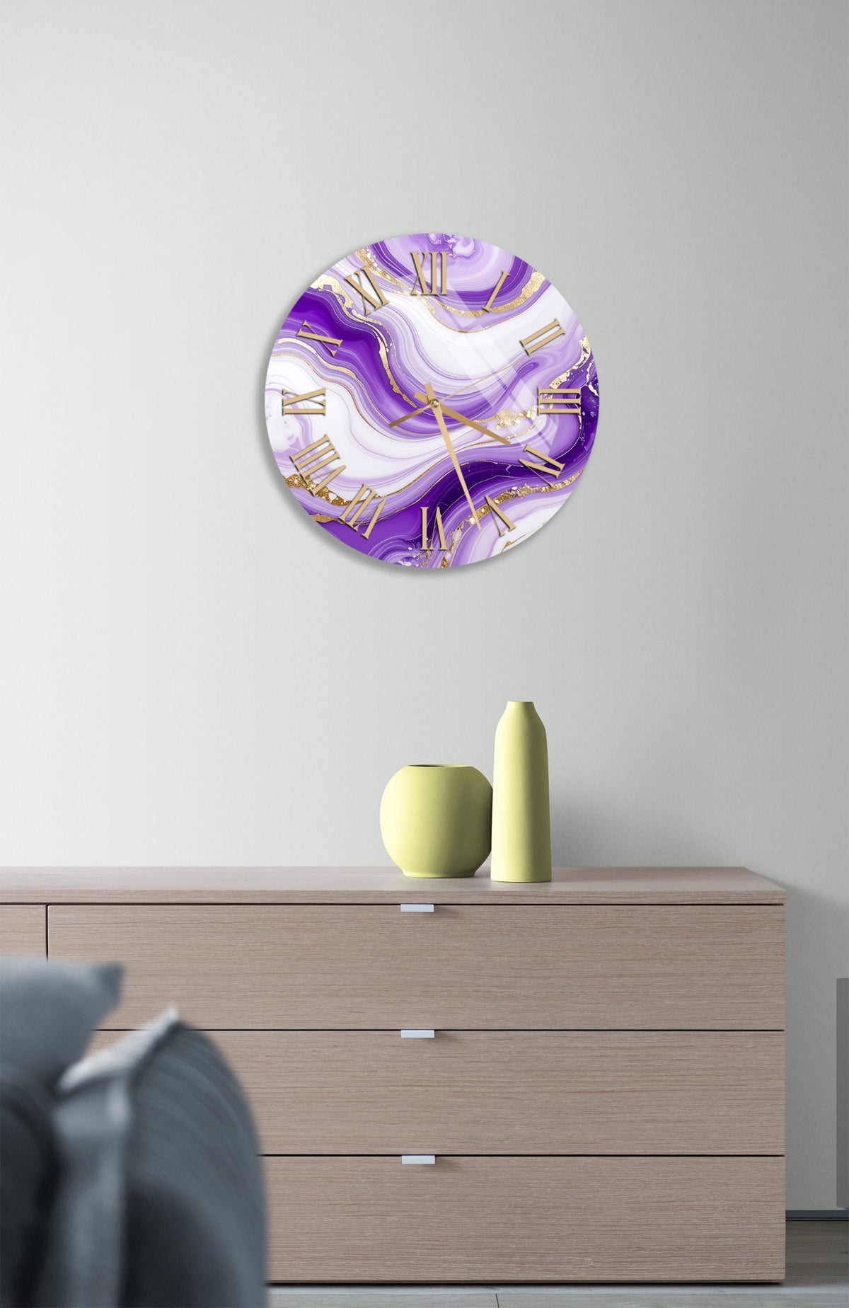 a modern interior design with a large round clock with a purple and white marble-like pattern on the wall above a wooden dresser. The dresser has three drawers and a small yellow vase on top.