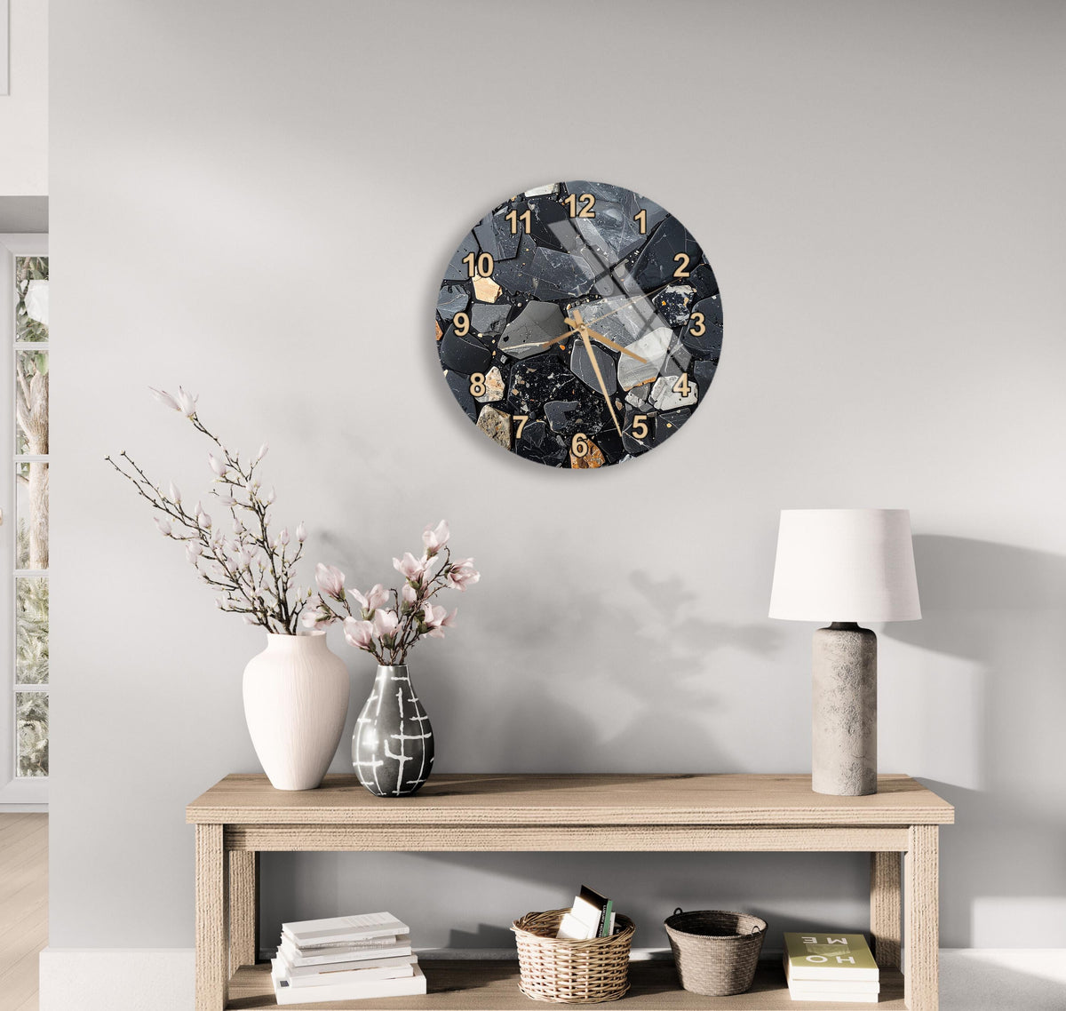 a modern clock with a black and white design, mounted on a wall above a wooden table with various decorative items, including a vase with flowers and a lamp.