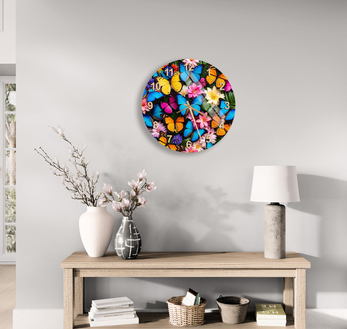 a round clock with a vibrant butterfly design on a white wall above a wooden table with a lamp, a vase with flowers, and some books.