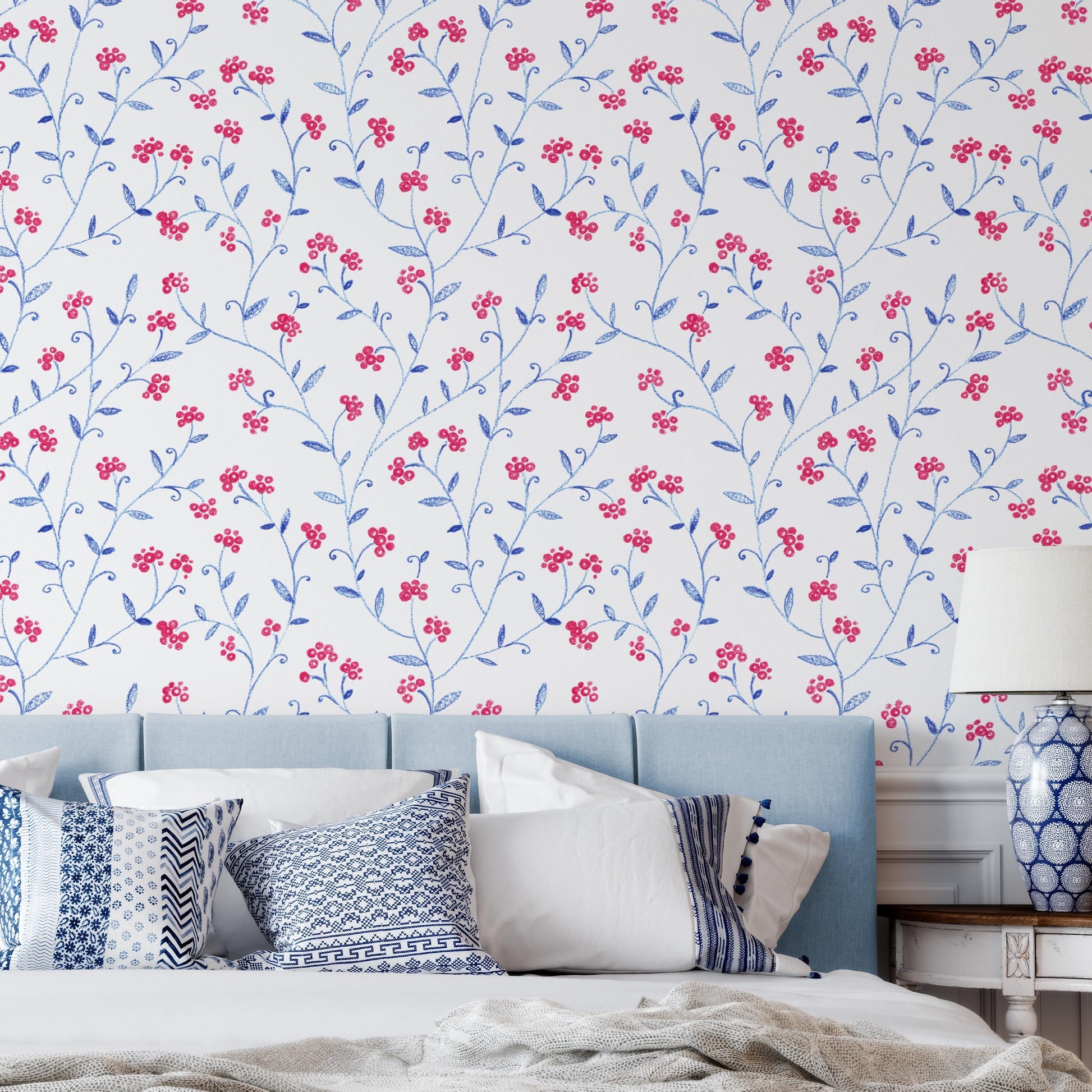 a bedroom with a blue headboard and a floral wallpaper pattern. The room has a white bed with blue and white patterned pillows, and a blue and white patterned lamp on a side table.